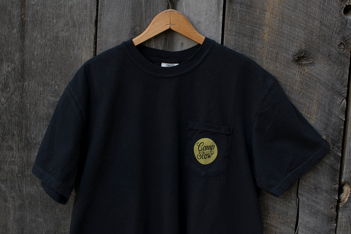 Black t-shirt with a green 'Camp and Go Slow' logo on a wooden hanger against a wooden background.