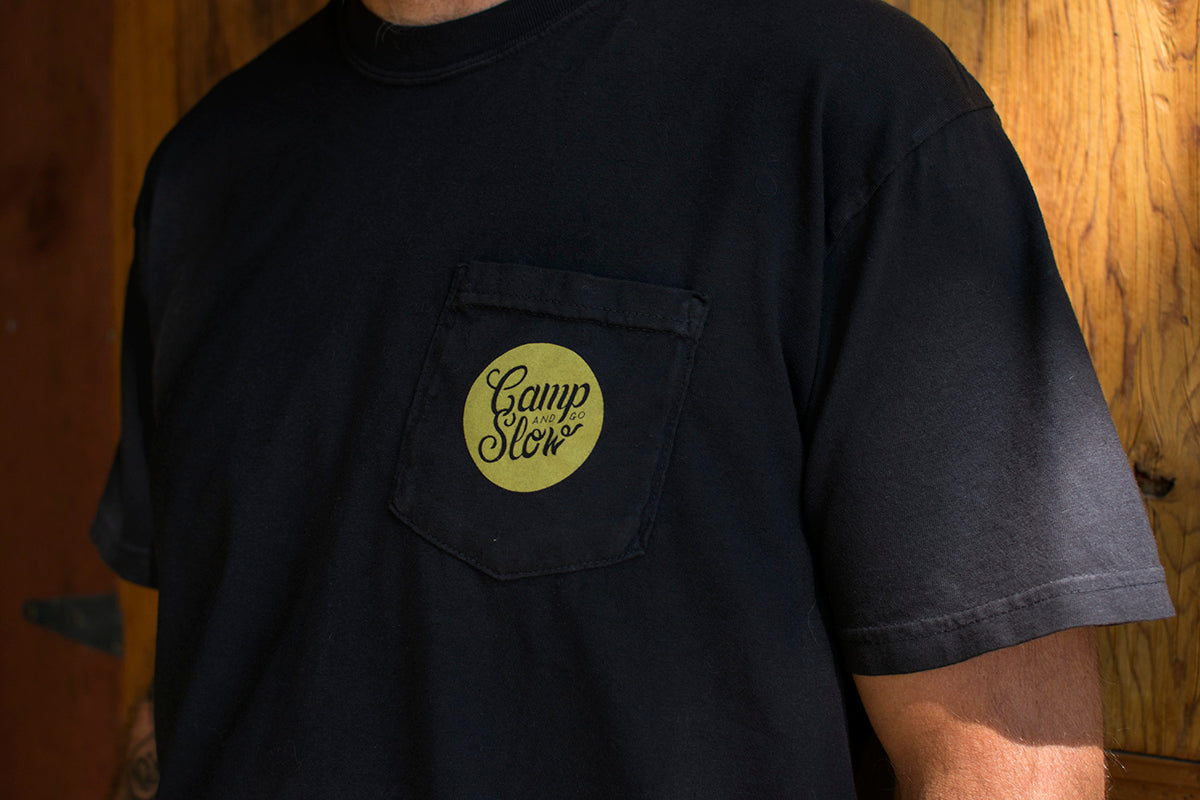 Black t-shirt with a green 'Camp and Go Slow' logo against a wooden background.