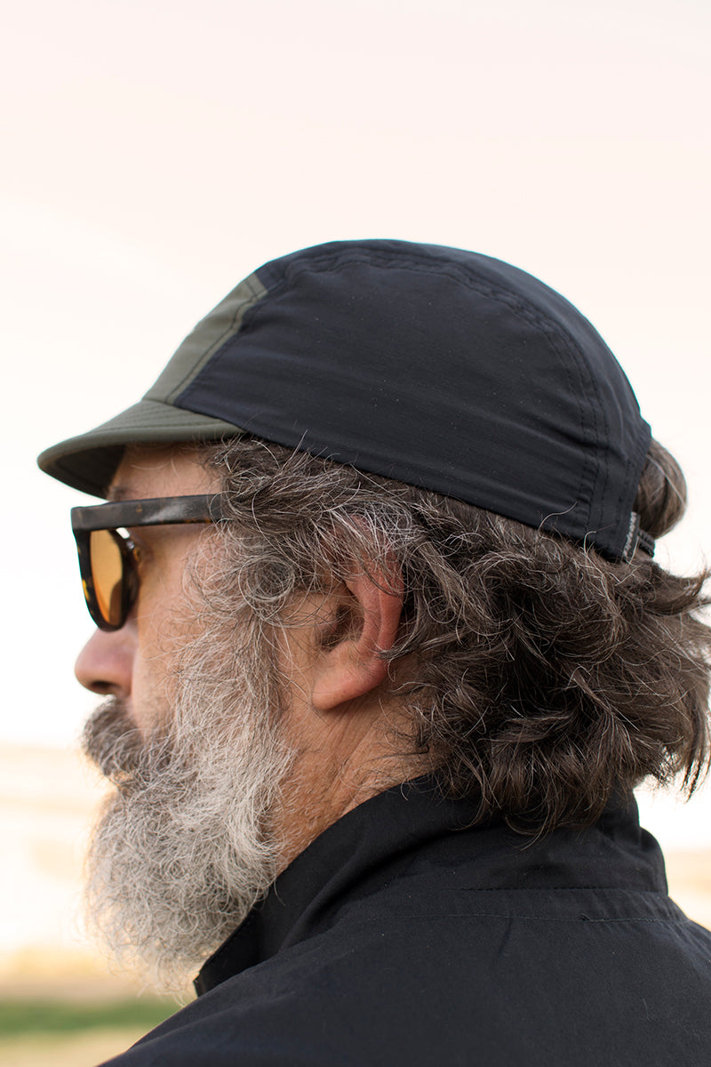 Man wearing a black and olive five panel cap and sunglasses with a blurred background