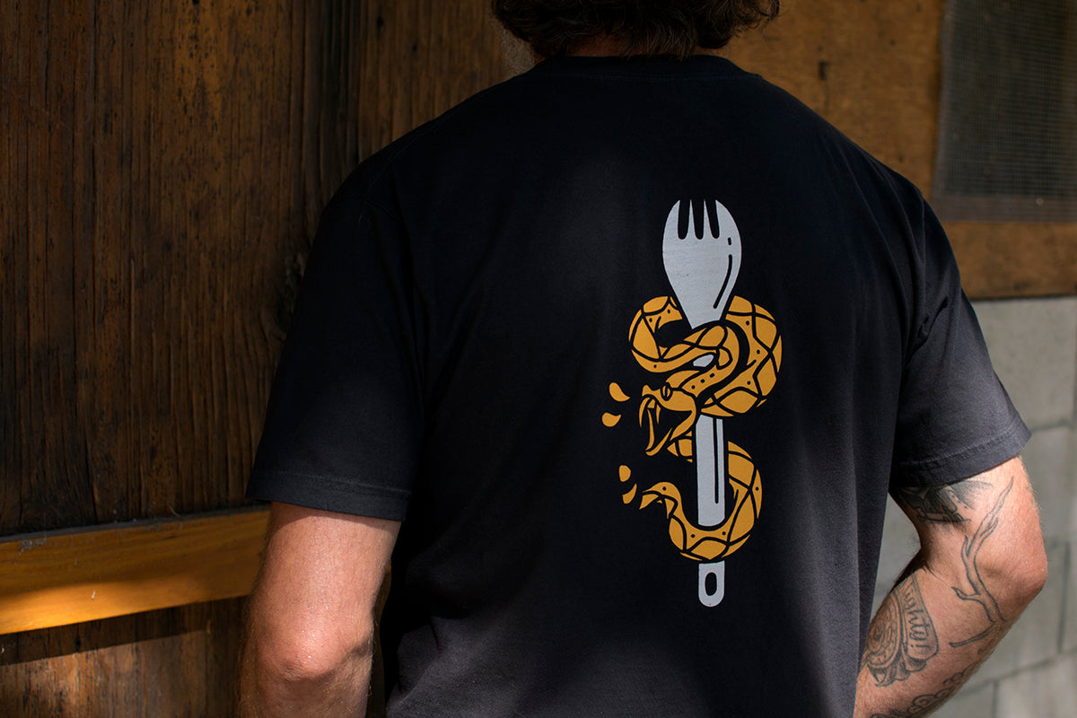 Person wearing a black t-shirt with a Western Rattler snake and spork design on the back, standing against a wooden wall.