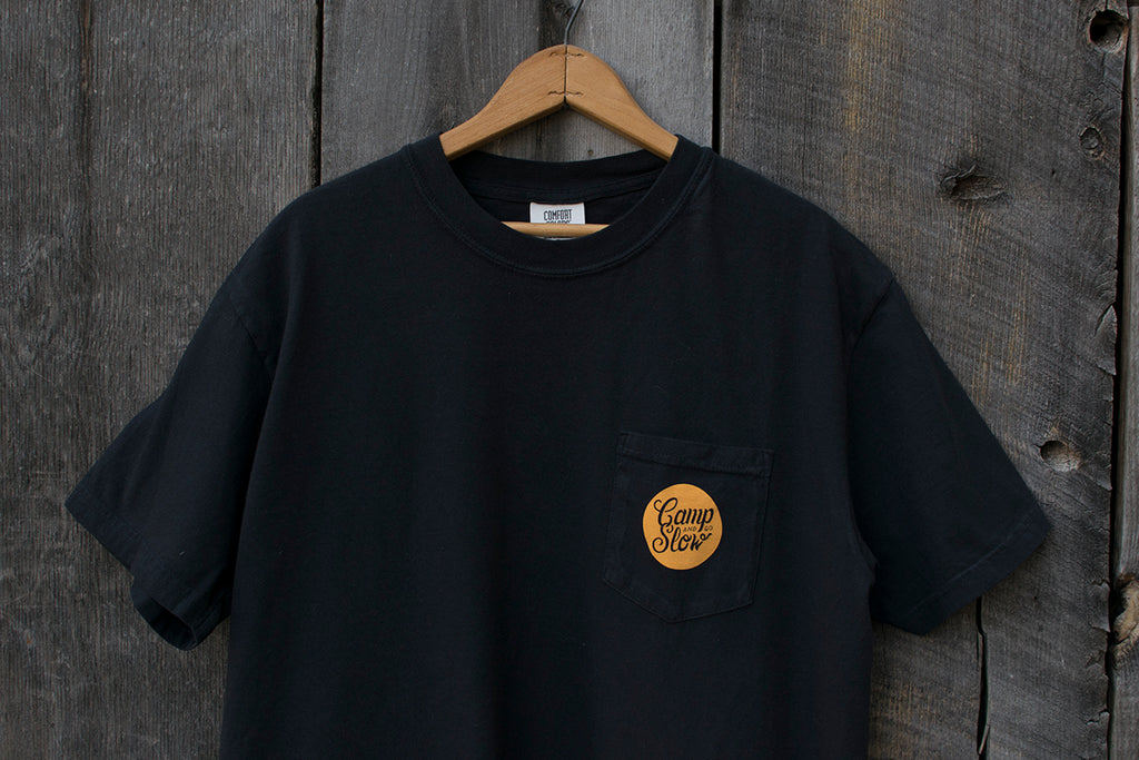 Black t-shirt with an orange 'Camp and Go Slow' logo on a wooden hanger against a wooden background