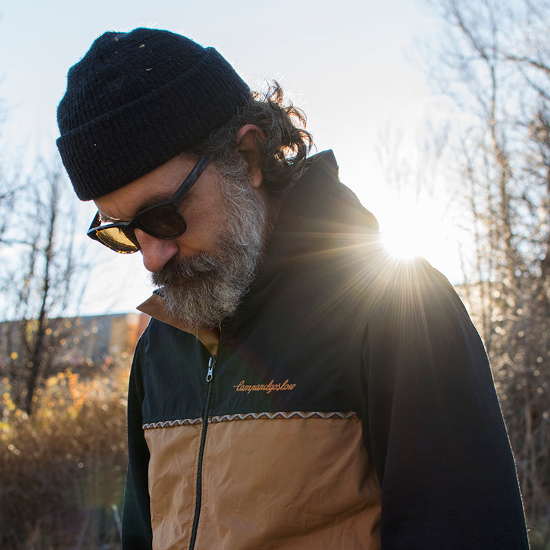 Man wearing a black beanie, sunglasses, and a Campandgoslow vest with a sunlit background